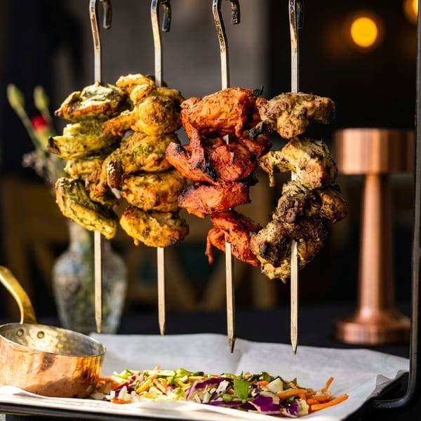Four skewers of colorful chicken tikka, with a side salad and copper pot, on a black table.