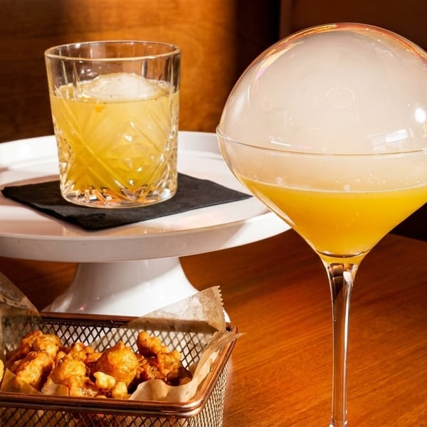 Two cocktails, one with a smoke bubble, and a basket of fried snacks on a wooden table.
