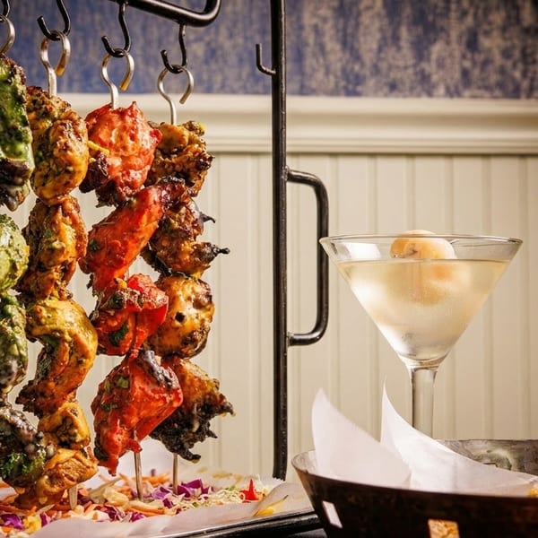 Colorful chicken skewers hanging from a rack, with a martini and bread basket on the right.