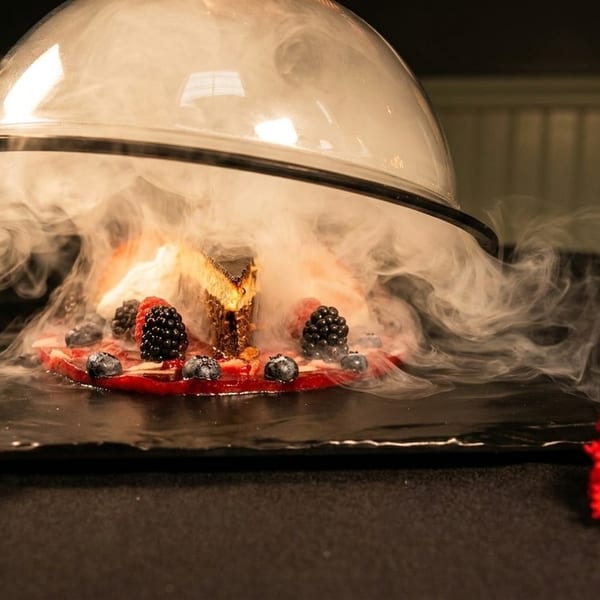 Cheesecake slice with berries and sauce, covered by a cloche emitting smoke, next to a red flower.