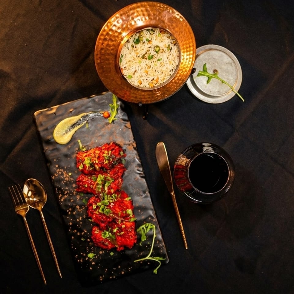 Overhead view of a black platter with red chicken, a copper bowl of rice, and a drink.